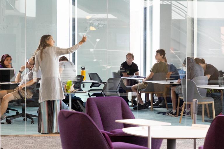 Students engage in a collaborative group discussion guided by an instructor in a glass-walled study room at the Learning Commons.